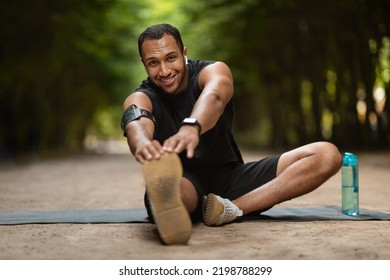 Positive Handsome Muscular Millennial Black Man Stretching In Public Park, Sitting On Yoga Mat, Reaching His Foot And Smiling, Enjoying Outdoor Workout, Copy Space. Sports Lifestyle And Millennials