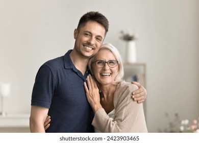 Positive handsome son man and happy blonde mature mother hugging at home with love, looking at camera with toothy smiles, laughing, standing close for family portrait - Powered by Shutterstock