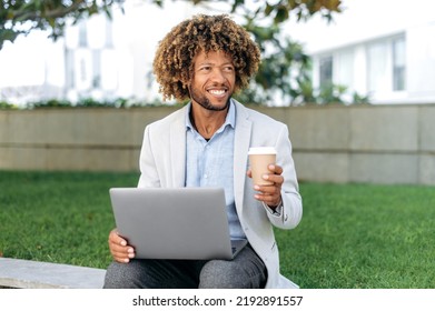 Positive Handsome Successful Brazilian Or Spanish Man, Company Employee, Entrepreneur, Sits On Outdoors With Laptop In His Hands, Holds A Glass Of Coffee In Hand, Pauses At Work, Looks Away, Smiles