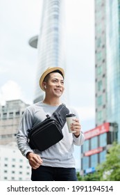 Positive Handsome Young Asian Man In Sun Hat And Wireless Earphones Holding Coffee Cup And Walking In Big City