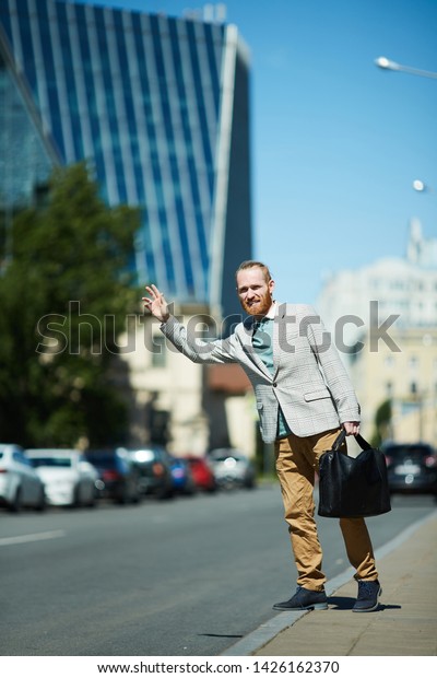 Positive handsome young\
businessman with briefcase waving hand while catching taxi on\
street