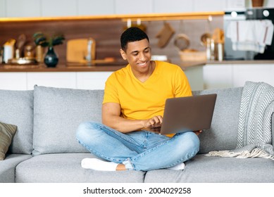 Positive Handsome Young Latino Man In Casual Clothes, Sitting On The Couch At Home In The Living Room, Using A Laptop, Browsing The Internet, Chatting On Social Networks, Looking At Screen, Smiles