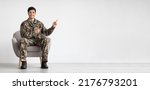 Positive handsome young soldier sitting in armchair and pointing with both hands at copy space for text or advertisement over white studio background, panorama, full length shot