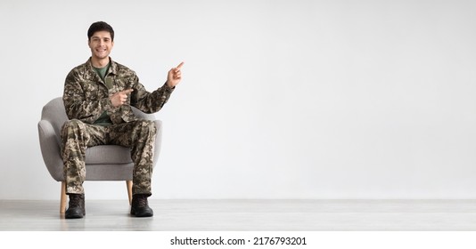 Positive handsome young soldier sitting in armchair and pointing with both hands at copy space for text or advertisement over white studio background, panorama, full length shot - Powered by Shutterstock