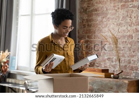 Positive happy African student girl unpacking box with books, receiving parcel with purchase from bookstore, opening cardboard package, paper container, moving, relocating in new home