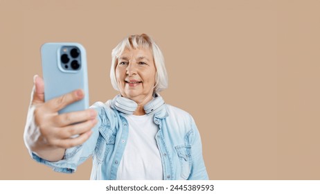 Positive happy elderly woman wearing casual clothes poses for selfie holding smartphone in one hand. Active old age concept - Powered by Shutterstock