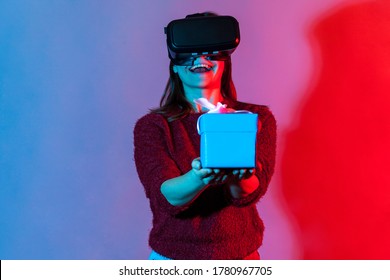 Positive Happy Girl In VR Virtual Reality Glasses Holding Wrapped Gift Box And Smiling Joyfully, Giving Christmas Present To Camera, Celebrating Holidays Birthday. Colorful Neon Light Studio Shot