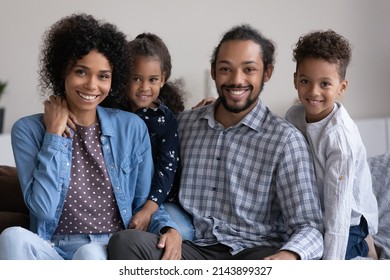 Positive happy young African family couple and two cute little sibling kids home head shot portrait. Black parents and preschool children sitting on couch, looking at camera, smiling