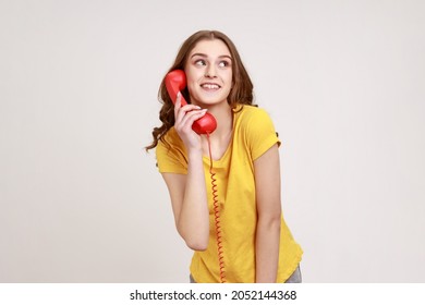 Positive happy young female with brown hair in yellow t-shirt talking landline telephone holding in hand handset, looking at away with dreamy look. Indoor studio shot isolated on gray background.