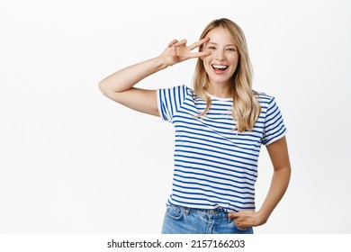 Positive And Happy Young Woman, 25 Years Old, Laughing And Smiling, Showing V-sign, Peace Gesture Near Face, Standing Over White Background