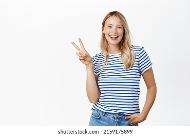 Positive And Happy Young Woman, 25 Years Old, Laughing And Smiling, Showing V-sign, Peace Gesture Near Face, Standing Over White Background