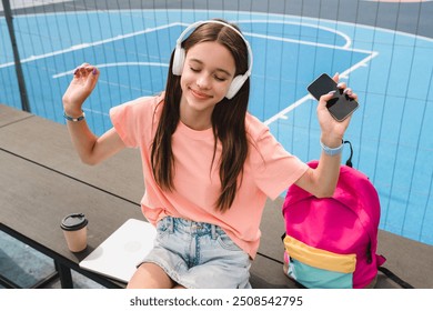 Positive high school girl teenager pupil college student in casual clothes with bag wireless headphone holding using mobile phone listening enjoying music dancing sitting in sport court yard outside - Powered by Shutterstock