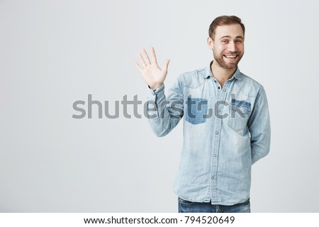Positive human emotions, facial expressions, feelings, attitude and reaction. Friendly-looking polite young European man dressed in denim shirt and jeans saying hi, waving with his hand