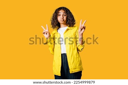 Positive human emotions. Headshot of happy emotional teenage girl with bob curly haircut with hands making peace sign, wearing bright makeup. Blue background. Wearing bright yellow cotton jachet.