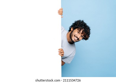 Positive Indian or Arabian guy, in casual t-shirt, peeking out from behind advertisement whiteboard, demonstrating blank copy space for your text or design, isolated blue background, mock-up concept