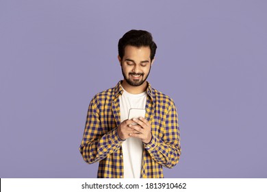 Positive Indian Man Looking At Mobile Phone Screen And Smiling On Violet Background