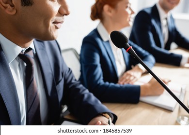 Positive Indian Politician Sitting Near Microphone With Blurred Colleagues On Background