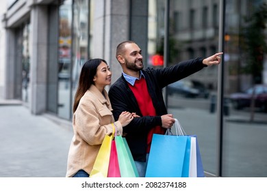 Positive Interracial Couple Walking Along Street With Paper Bags, Pointing At New Store Window, Shopping Together Outdoors. Young Diverse Family Enjoying Seasonal Sales, Buying Goods On Discount