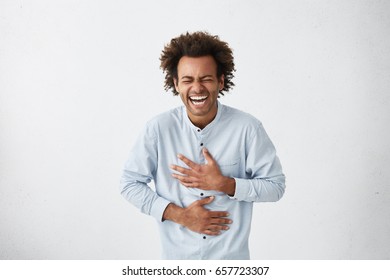 Positive and joyful Afro American male with fine crop of hair bursting into laughing holding his hands on stomach can`t stopping laughing after hearing funny anecdote. Positive emotions and humour - Shutterstock ID 657723307