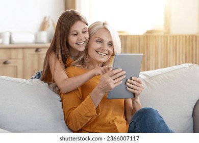 Positive joyful attractive blonde elderly woman 60s having fun with her grandchild pretty teen aged girl at home, sit on couch in living room, using digital tablet, play mobile game, using nice app - Powered by Shutterstock