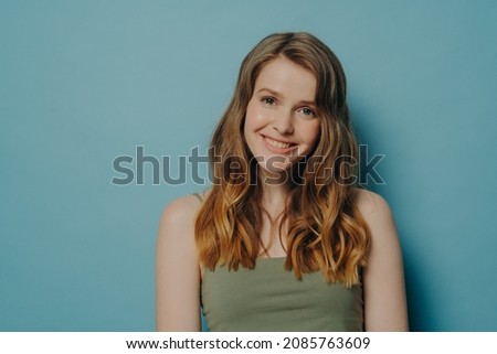 Positive joyful teen girl in top tank demonstrating optimism and happiness while standing isolated over blue studio background with copy space for text, pleased happy female looking at camera