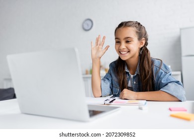 Positive Kid Waving Hand During Video Chat On Laptop Near Blurred Sticky Notes At Home