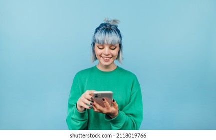 Positive lady with blue hair and green sweatshirt enjoying smartphone on blue background with smile on face looking at screen. - Powered by Shutterstock