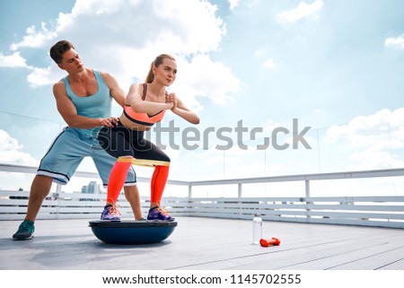 Positive lady is doing fitness with male instructor on terrace of high building. She is using both BOSU platform and resistance band during work out with dumbbells. Copy space in right side