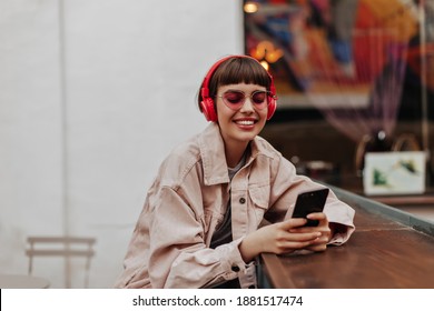 Positive lady with red headphones smiling in cafe. Modern girl in beige denim jacket and pink sunglasses listening to music and holding phone outside..