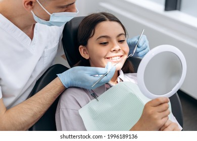 Positive little girl holding a mirror, sitting in a dental chair. The dentist checks the teeth of a young patient - Powered by Shutterstock