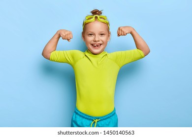 Positive Little Girl Spends Leisure Time In Water Park, Raises Arms And Shows Muscles, Ready For Swim And Dive, Poses Over Blue Background, Wears Goggles And Swimsuit. Small Kid Has Healthy Lifestyle