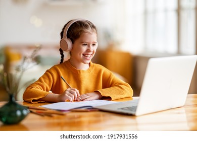 Positives kleines Schulmädchen in Kopfhörern, das mit dem Tutor kommuniziert und während des Online-Unterrichts auf Laptop zu Hause Notizen in das Copybook macht