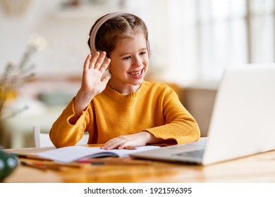 Positive little schoolgirl in headphones communicating with tutor waving hands and greeting tutor during video call via laptop while having online lesson at home - Shutterstock ID 1921596395