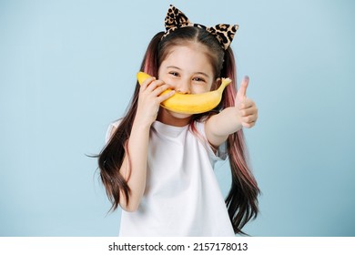 Positive Looking Tween Girl With Cat Ears Holding Banana Over Her Mouth As A Smile. She Has Dyed Hair Strands. Posing Over Blue Background.