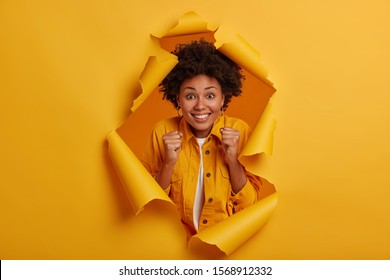 Positive lucky woman with Afro hairstyle, raises clenched fists, smiles broadly, poses in ripped hole background, enjoys perfect day, anticipates for something awesome, screams from delight.