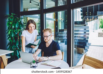 Positive male copywriter posting article on content website while female friend standing near with textbook in hand and giving consultancy about 4g connection on laptop computer,concept of cooperation