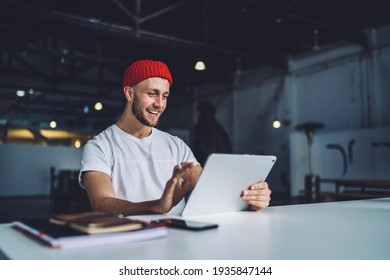 Positive male editor in trendy cap sitting at desktop with modern touch pad wathcing video before online montage via application, happy man in stylish outfit reading good news on content website