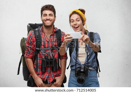 Positive male and female adventurers having happy expressions posing in camera of their modern smart phone isolated over white wall with copy space. Cheerful friends having hiking trip together
