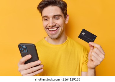 Positive male shopper pays online via smartphone holds credit card downloads ebanking application satisfied with easy quick safe way to pay provides distant payment dressed in casual t shirt