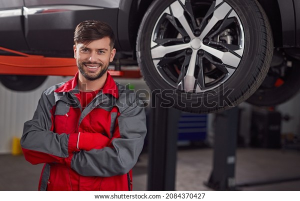 Positive male technician crossing arms and looking\
at camera with smile while standing near modern vehicle on\
hydraulic lift in\
garage