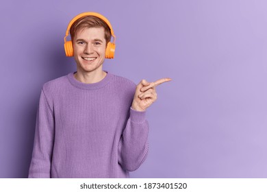 Positive Male Teenager With Blue Eyes And Happy Smile Wears Casual Purple Sweater Listens Music In In Stereo Headphones Points Away On Copy Space Advertises Something Over Copy Space. Youth Hobby