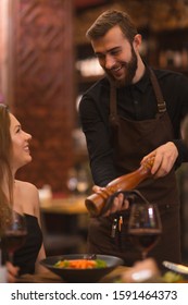 Positive Male Waiter In Black Shirt And Apron Seasoning Meals Of Client In Restaurant. Handsome Man Talking With Woman, Joking And Laughing While Servicing And Doing Job. Concept Of Dinner.