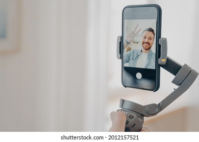 Positive man blogger holding gimbal stabilizer with smartphone and trying to make selfie pictures and live video while spending time at home in living room. Vlog and video blogging concept
