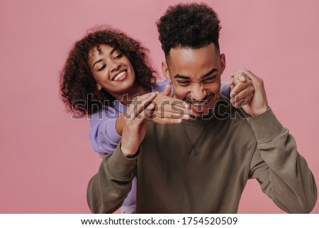 Positive man in brown outfit laughing and holding his girlfriends hands. Cool guy and curly brunette woman dressed in purple shirt smiling on pink background