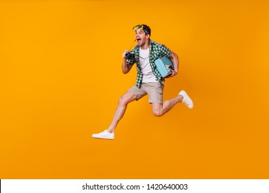 Positive Man With Diving Mask On His Head Takes Pictures With Retro Camera. Guy In Shorts And Green Shirt Jumping With Suitcase On Orange Background