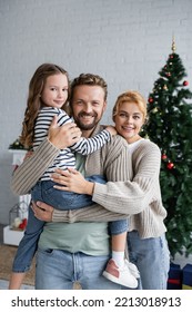 Positive Man Holding Daughter Near Wife In Cardigan And Blurred Christmas Tree At Home