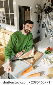 Positive man in jumper looking at camera while sitting near fresh food in modern kitchen at home - Shutterstock ID 2395465771
