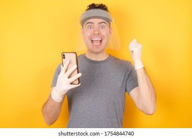 Positive Man Listens Audio Record With High Volume, Holds Modern Cell Phone Connected To Headphones, Clenches Fist From Good Emotions, Exclaims With Joy, Wears Casual Green Tshirt