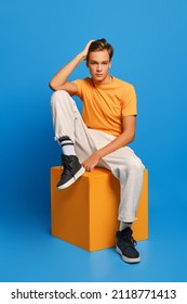 Positive man in orange undershirt and white sweatpants sits on wooden cube over blue studio background