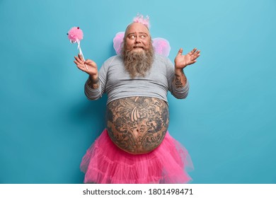 Positive man in princess outfit, wears pleated rosy skirt, wings and undersized t shirt, holds magic wand, prepares for carnival or costume party, has big tattooed belly, waits for miracle happened